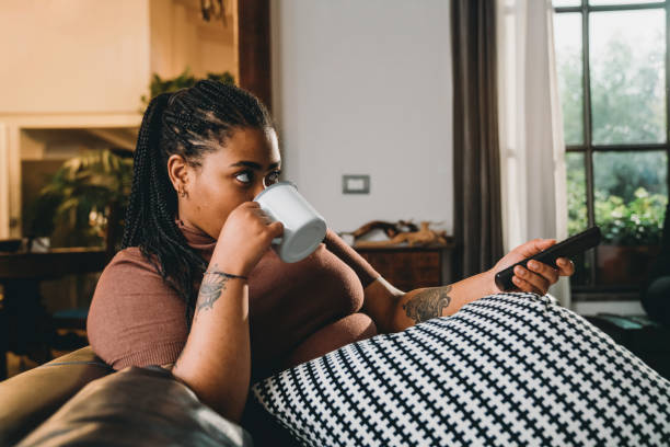 Mixed race woman is watching TV and drinking a coffee on the sofa Mixed race woman is watching TV and drinking a coffee on the sofa. She's relaxing herself at home. streaming service stock pictures, royalty-free photos & images