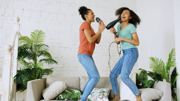 Mixed race young funny girls dance singing with hairdryer and comb jumping on sofa. Sisters having fun leisure in living room at home concept stock photo