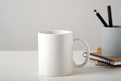 Mockup of a white mug on a table in a minimalist interior, business stationery and notepads. Template, layout for your design, advertising, logo with copy space. Cup on light gray background.
