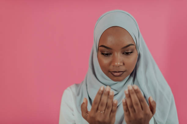 Modern African Muslim woman makes traditional prayer to God, keeps hands in praying gesture, wears traditional white clothes, has serious facial expression, isolated over plastic pink background stock photo
