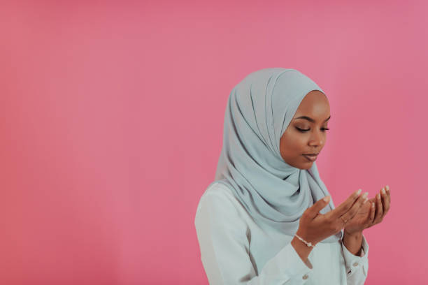 Modern African Muslim woman makes traditional prayer to God, keeps hands in praying gesture, wears traditional white clothes, has serious facial expression, isolated over plastic pink background stock photo