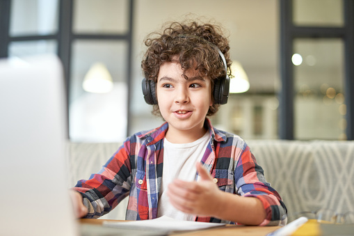 Portrait of joyful latin american little boy wearing headphones, looking at the screen of a laptop while having online lesson at home. Online education, technology, homeschooling concept