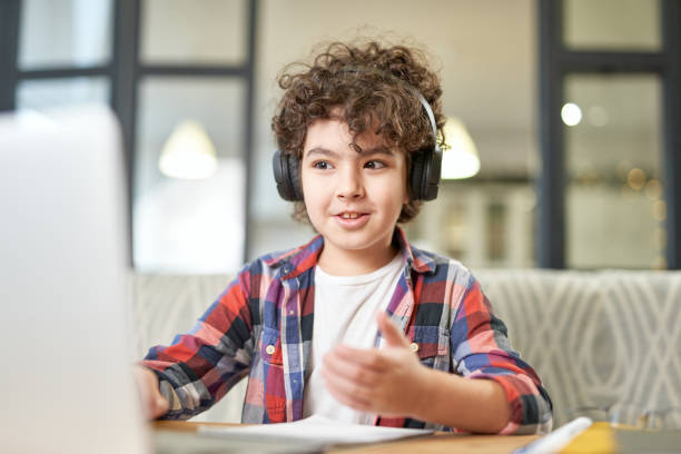 Modern approach to learning. Portrait of joyful latin american little boy wearing headphones, looking at the screen of a laptop while having online lesson at home stock photo