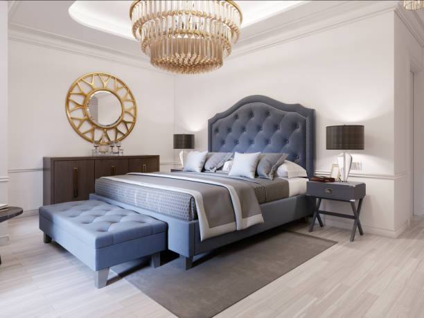 Modern bed in classic blue style with bedside table and lamp. Large glass chandelier over. A dresser with a decor and a golden mirror above. Modern bedroom. stock photo