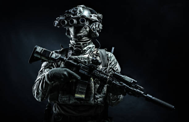 Modern combatant wearing night vision device black background Army infantry in battle uniform, armed assault rifle with laser sight and silencer, standing in darkness, looking through night vision goggles, low key half length, studio portrait on black background special forces stock pictures, royalty-free photos & images