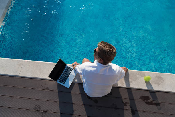 A modern elderly man enjoys the pool while working on his laptop next to a modern luxury house. Selective focus stock photo