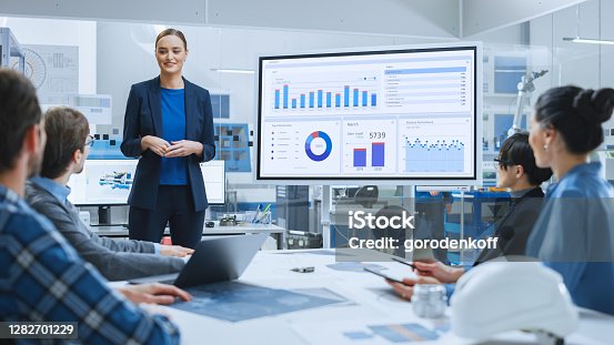 istock Modern Industrial Factory Meeting: Confident Female Engineer Uses Interactive Whiteboard, Makes Report to a Group of Engineers, Managers Talks and Shows Statistics, Growth and Analysis Information 1282701229