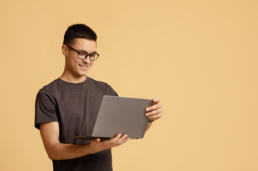 Modern remote communication online, stay at home, during covid-19 lockdown. Smiling millennial guy student with glasses looks at laptop and works on project, isolated on beige background, studio shot