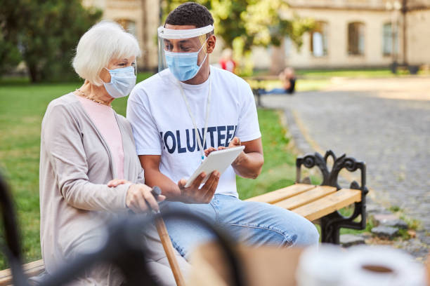 Modern technologies becoming easier for elderly people Young male volunteer wearing a protective shield while sitting on a bench with an aged woman in medical mask and showing a modern tablet volunteer stock pictures, royalty-free photos & images