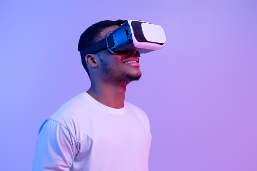 Modern technologies for entertainment. Excited black man wearing vr glasses looking aside at copy space, african guy experiencing virtual reality, standing on purple background in neon lighting