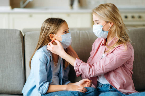 Mom and daughter in masks at home. A caring mother in a protective mask puts a medical mask to her daughter to protect from a virus or flu while sitting on a couch at their home, healthcare concept stock photo