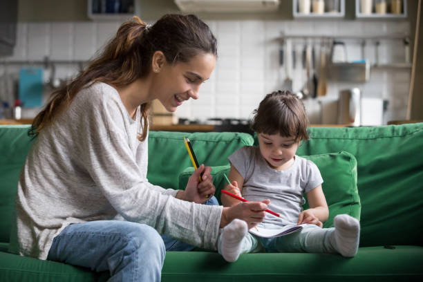Mom and kid girl drawing with colored pencils at home Smiling baby sitter and preschool kid girl drawing with colored pencils sitting on sofa together, single mother and child daughter playing having fun, creative family activities at home concept Nanny stock pictures, royalty-free photos & images