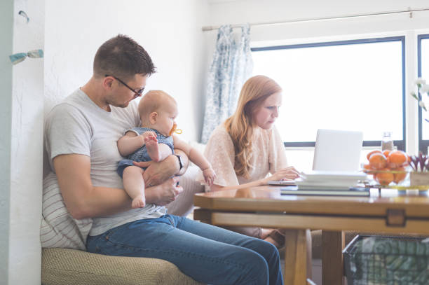 Mom working at her computer while dad holds their baby daughter Dad holds their daughter at the kitchen table while Mom checks her email, minus baby, on her laptop next to him. gender stereotypes stock pictures, royalty-free photos & images