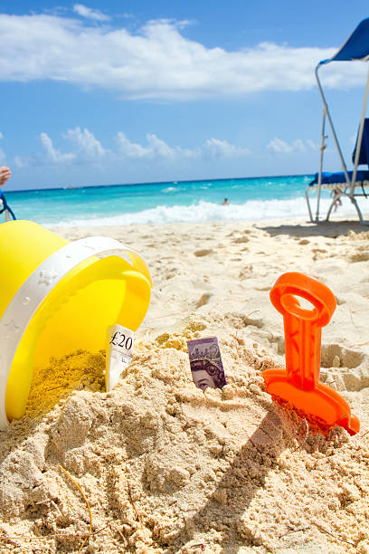 money in sand on tropical beach indicating holiday cost English bank notes sticking out of the sand next to an upturned bucket on a tropical beach in Barbados on a glorious sunny day with blue sky. Foot of person relaxing on a lounger and blurred image of person in the sea in background. human feet buried in sand. summer beach stock pictures, royalty-free photos & images