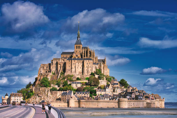 Mont-Saint-Michel / France Normandy, France, August 29, 2019: Looking at the magestic Mont Saint-Michel  island during low tide, it makes the city look like it was built on dry sand.  Le Mont-Saint-Michel or in english Saint Michael's Mount is a tidal island and mainland commune in Normandy, France. The island lies approximately one kilometre (0.6 miles) off the country's north-western coast, at the mouth of the Couesnon River near Avranches and is 7 hectares (17 acres) in area. The mainland part of the commune is 393 hectares (971 acres) in area so that the total surface of the commune is 400 hectares (988 acres). As of 2017, the island has a population of 30. 
As of 2017, the island has a population of 30. Mont-Saint-Michel and its bay are on the UNESCO list of World Heritage Sites. It is visited by more than 3 million people each year. Over 60 buildings within the commune are protected in France as monuments historiques. manche stock pictures, royalty-free photos & images