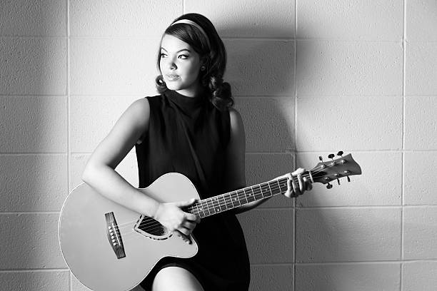 Moody B&W of young 60s styled woman with guitar. Horizontal studio shot on white brick background of mixed race teen in 60s style hair and minidress playing guitar and looking away. girls in very short dresses stock pictures, royalty-free photos & images