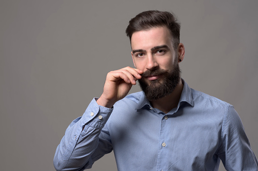 Moody portrait of young stylish bearded man twirling mustache and looking at camera over gray studio background with copyspace.