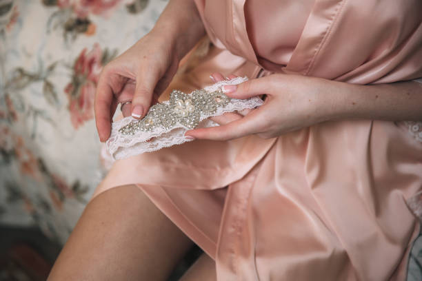 Morning of the bride. Close-up of young bride in pink dressing-gown putting on white garter at the wedding day. Hands of the girl with accessories stock photo