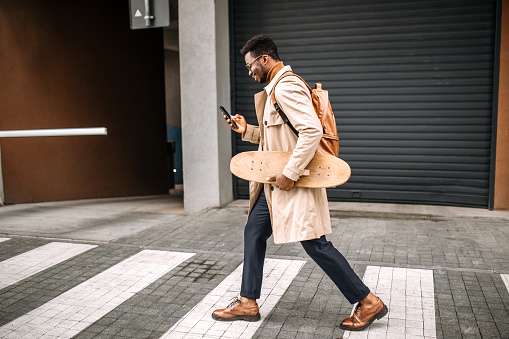 Young man with backpack walking on his way to work, carrying skateboard, using smart phone