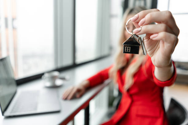 Mortgage concept. Woman in red coral business suit holding key with house shaped keychain. Modern light lobby interior. Real estate, hypothec, moving home or renting property. stock photo