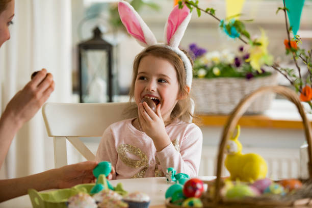 Mother and daughter celebrating Easter, eating chocolate eggs. Happy family holiday. Cute little girl in bunny ears laughing, smiling and having fun. stock photo