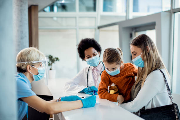 Mother and daughter checking in at medical clinic during coronavirus pandemic. Little girl and her mother talking to a nurse at reception desk in the hospital during COVID-19 pandemic. hotel reception photos stock pictures, royalty-free photos & images