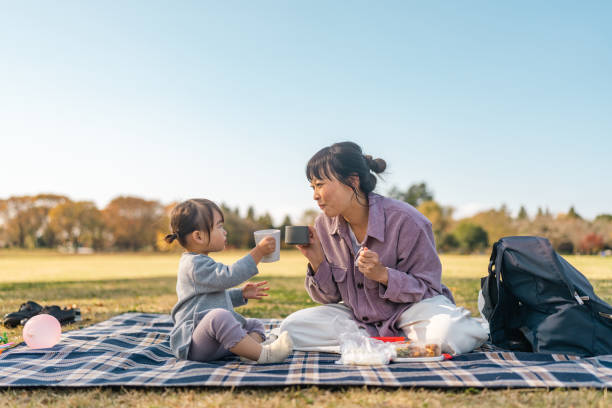 Mother and daughter enjoying having picnic together in public park on warm sunny day stock photo
