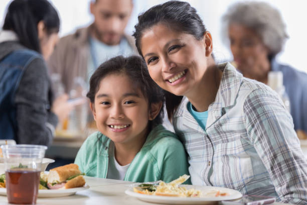 Mother and daughter have lunch in soup kitchen Mid adult woman and her elementary age daughter have lunch in a soup kitchen. People and volunteers are in the background. filipino family stock pictures, royalty-free photos & images