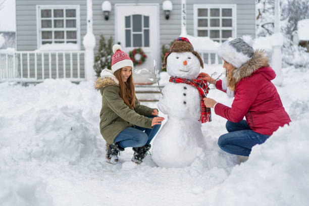 Mother and daughter making snowman in front of the house, enjoying winter fun together on a snowy winter day stock photo