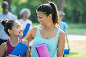 Mid adult Asian Filipino woman is smiling and has her arm around her elementary age daughter. Mother and child are attending an outdoor yoga class together in local park, and are holding exercise mats.