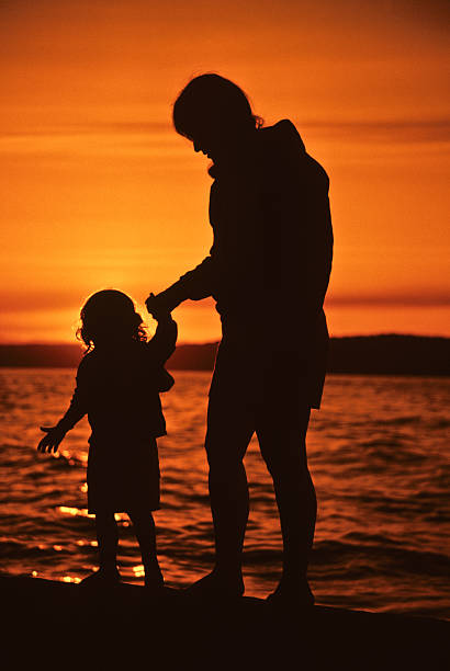Mother and Daughter, Sunset Silhouette Puget Sound is a special body of water that belongs exclusively to Washington State. From the saltwater marshland of Olympia and Nisqually to the windy Strait of Juan de Fuca and the San Juan Islands, Puget Sound exhibits a quiet beauty that is quintessentially Washington. Puget Sound and its adjacent waters form a vast inland extension to the Pacific Ocean. Its complex network of straits, bays, canals and inlets give Western Washington a distinctive personality. Puget Sound along with its surrounding mountains has a major effect on the region's temperate climate. The area was first visited by Europeans in 1792 with British explorer George Vancouver's expedition. Captain Vancouver gave the region many of the place names which are still in use today. Not only does Puget Sound have a rich history, it also has some of the greatest scenery in the country. Sunsets over Puget Sound can be especially beautiful and an inspiration to photographers. This silhouette picture of a mother and daughter was photographed as the sun set over Puget Sound from Dash Point State Park near Tacoma, Washington State, USA. jeff goulden puget sound stock pictures, royalty-free photos & images