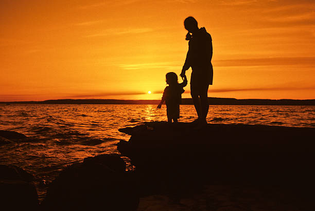 Mother and Daughter, Sunset Silhouette Puget Sound is a special body of water that belongs exclusively to Washington State. From the saltwater marshland of Olympia and Nisqually to the windy Strait of Juan de Fuca and the San Juan Islands, Puget Sound exhibits a quiet beauty that is quintessentially Washington. Puget Sound and its adjacent waters form a vast inland extension to the Pacific Ocean. Its complex network of straits, bays, canals and inlets give Western Washington a distinctive personality. Puget Sound along with its surrounding mountains has a major effect on the region's temperate climate. The area was first visited by Europeans in 1792 with British explorer George Vancouver's expedition. Captain Vancouver gave the region many of the place names which are still in use today. Not only does Puget Sound have a rich history, it also has some of the greatest scenery in the country. Sunsets over Puget Sound can be especially beautiful and an inspiration to photographers. This silhouette picture of a mother and daughter was photographed as the sun set over Puget Sound from Dash Point State Park near Tacoma, Washington State, USA. jeff goulden sunset stock pictures, royalty-free photos & images