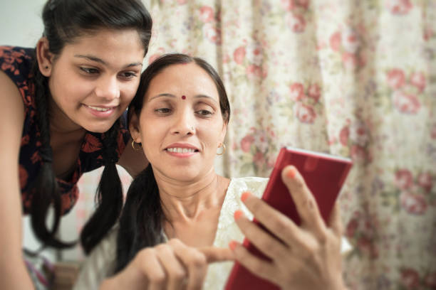 Mother and daughter using phablet together in domestic room. Indoor image of happy mature Asian women using phablet with her teenage daughter in her domestic room. Two people selective focus and horizontal composition. asian mother talking with daughter stock pictures, royalty-free photos & images