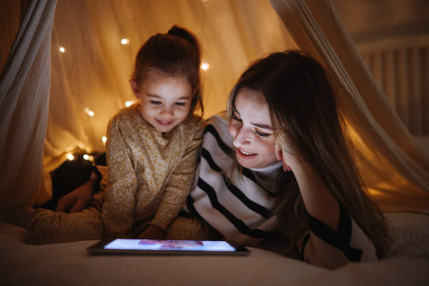 moeder en dochter kijken samen naar hun digitale tablet terwijl ze in bed onder de tent liggen.  - ledstrips stockfoto's en -beelden