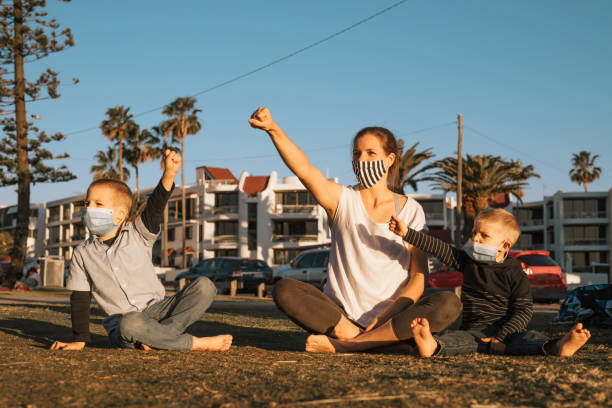 Mother and her two little sons are sitting on the grass wearing protective masks and holding arms with fists up. stock photo