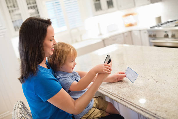Mother And Son Depositing Check Through Smart Phone Side view of mother and son depositing check through smart phone in kitchen bank deposit slip stock pictures, royalty-free photos & images