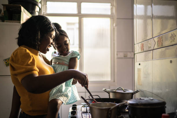 mother cooking at home while holding her daughter - black mother imagens e fotografias de stock