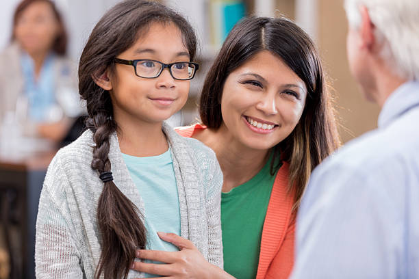 Mother discusses daughter's symptoms with male pediatrician Cheerful mother talks with senior doctor about her daughter's symptoms. The girl is smiling at the doctor. asian mother talking with daughter stock pictures, royalty-free photos & images