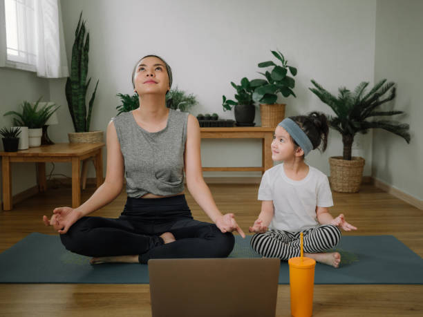 Mother doing on yoga mat with little daughter at home. Asian little girl and her mother enjoying free time at home in the living room, practicing yoga together. asian yoga pants stock pictures, royalty-free photos & images