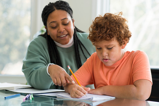 Beautiful mother is smiling gently as she is helping her elementary age son with his homework from school at the kitchen table. She is pointing at a mistake on the page and he is using a pencil to correct the work in the notebook.