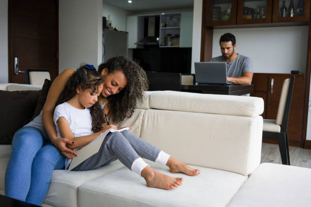 madre ayuda a su hija con la tarea mientras que papá está trabajando en el ordenador portátil - latin family fotografías e imágenes de stock