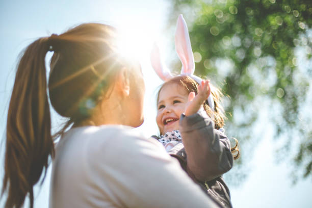 Mother holding child with bunny ears Cute 2 years old toddler girl playing with a basket full with Easter eggs and carrots outside under a big tree. She is happy and wearing Easter bunny ears headband easter stock pictures, royalty-free photos & images