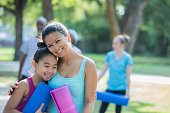 Mid adult Asian Filipino woman hugs her elementary age daughter as they attend an exercise class outdoors together in local park. Mother and child are holding yoga mats.