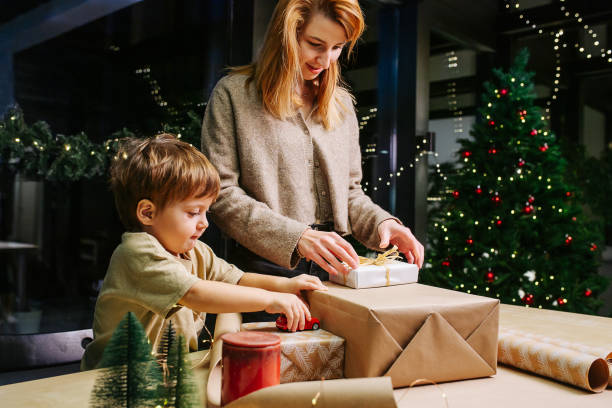 Mother prepares for christmas, wrapping presents in paper next to her son stock photo