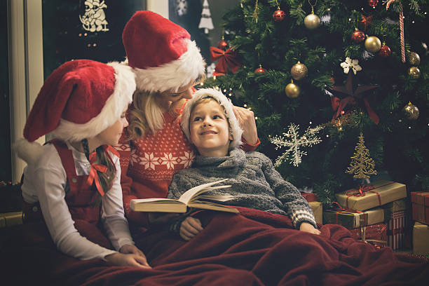 Mother reading a book to children during Christmas holidays Mother reading a book to children during Christmas holidays. Mother and children are wrapped in a blanket and sitting on the floor by the Christmas tree. christmas story telling stock pictures, royalty-free photos & images