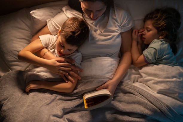 Mother reading stories to her kids before speeping while they lying in bed stock photo