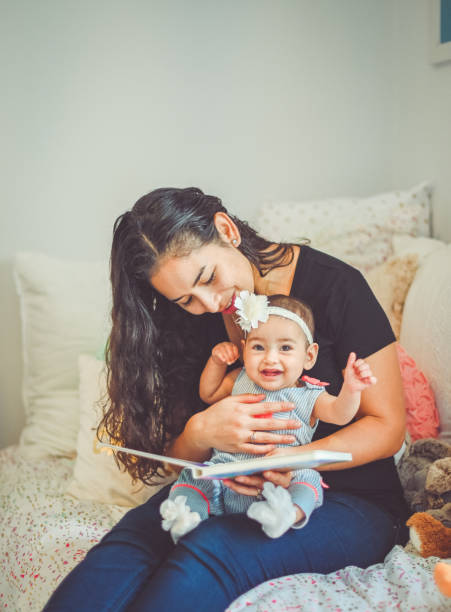 Mother reads book to sweet 6 month old baby Cute mother and daughter Latina pair read a book together at home in a sweet candid moment. Little girl is around 6 months old and her pretty mother sits on a bed reading with her cute puerto rican girls stock pictures, royalty-free photos & images