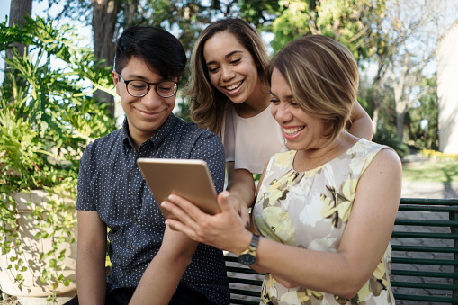 A latin mother holding and showing a digital tablet to her two young adult children outside and smiling.