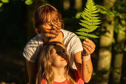 Mother, with long brown hair tied in a braid, showing her little daughter, with blonde hair, a beautiful green leaf in the forest, holding it against the sunlight while looking at it, leaf is casts a shadow on the face of the mother, forest in the background, front view, focus on forefront, horizontal