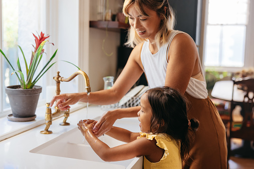 Mother teaching her daughter to wash her hands with soap and running water. Loving mom following precautionary measures at home. Mother and daughter standing by the sink in the kitchen.
